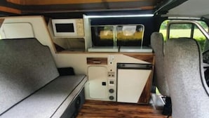 Interior compacto de una furgoneta con kitchenette, una televisión pequeña y una zona de estar acolchada.