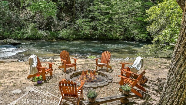 A circular seating area with wooden Adirondack chairs around a fire pit, set on a gravel surface with a river in the background. - Trout Haven | No Pet Fees | Sleeps 6 | Creekfront | Outdoor pavillion w/ TV (Ellijay)