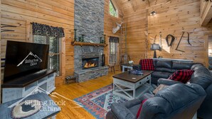 A log cabin interior with a stone fireplace, leather sofa, and a television on a wooden stand. - Trout Haven | No Pet Fees | Sleeps 6 | Creekfront | Outdoor pavillion w/ TV (Ellijay)