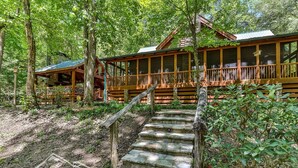 A cabin with a wooden deck, stone steps, and a forested backdrop. - Trout Haven | No Pet Fees | Sleeps 6 | Creekfront | Outdoor pavillion w/ TV (Ellijay)
