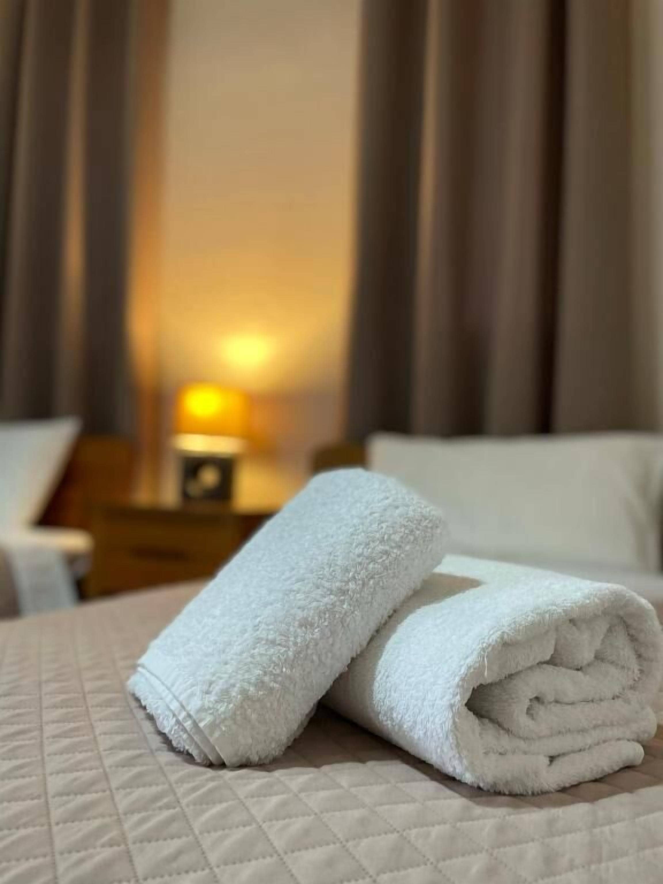 A neatly made bed with a quilted cover, a folded white towel, and a bedside table with a lamp.