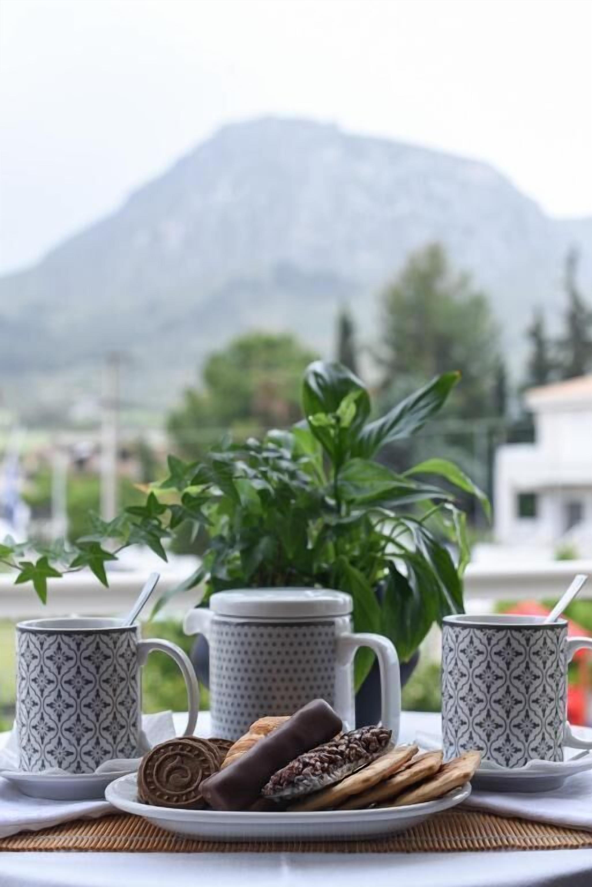 A table with a plate of cookies and two patterned mugs, a teapot, and a cup with a straw, set against a mountain backdrop.