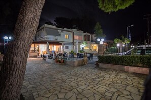 A stone-paved outdoor area with tables and chairs, a building with multiple floors and balconies, and a flag in the background. - Vasilios Marinos Rooms (Corinth)