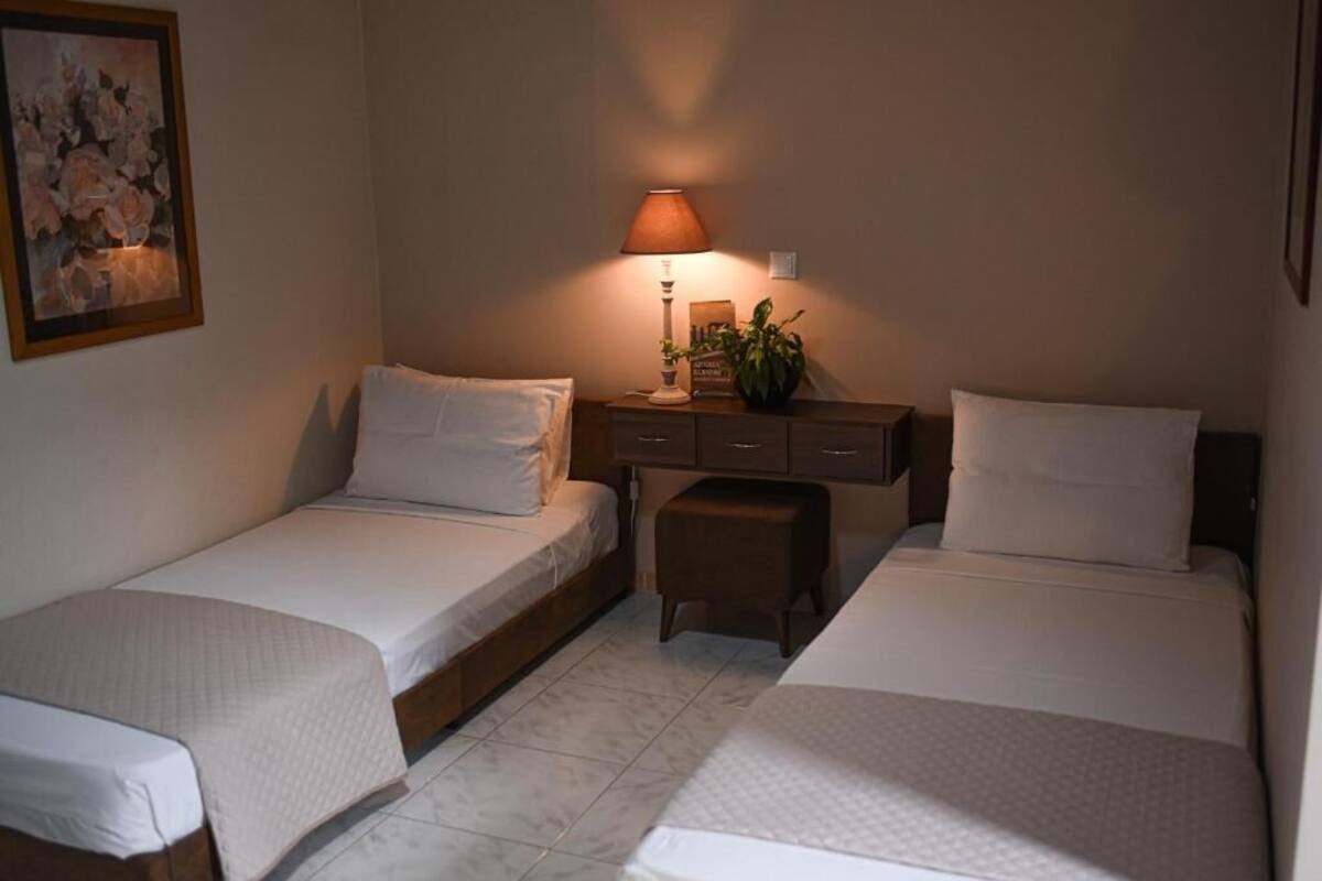 Ein kleiner, gut beleuchteter Raum mit zwei Einzelbetten, einem Nachttisch mit Lampe, einem kleinen Tisch mit Pflanze und einem gerahmten Bild an der Wand.