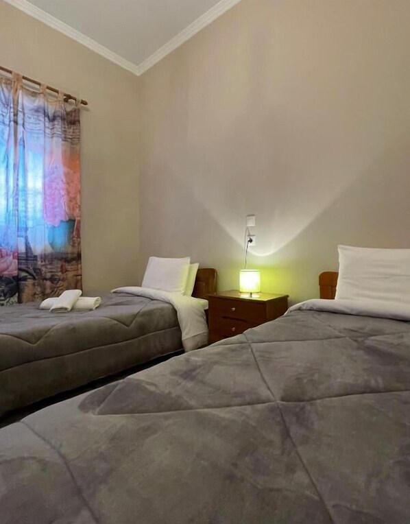 Habitación de hotel con dos camas, una mesita de noche con lámpara y una ventana con cortinas.