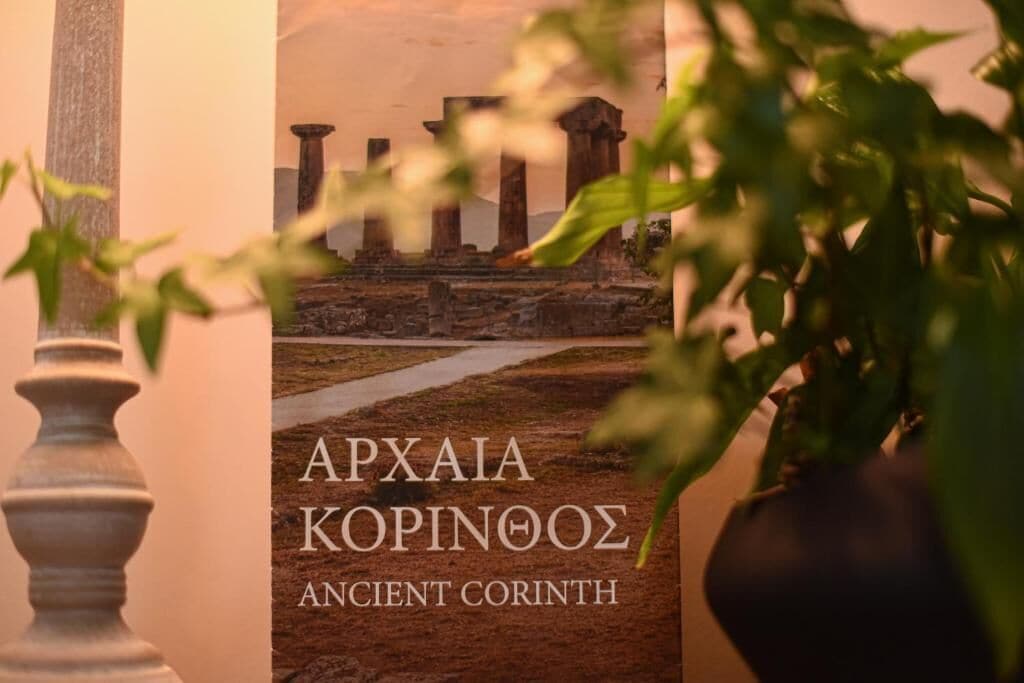 A book titled 'APXAIA KOPINOΩΣ ANCIENT CORINTH' with a background of ancient ruins and a pathway.