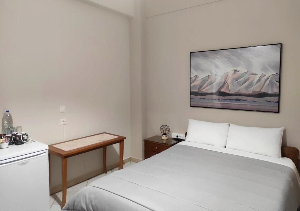 A hotel room with a bed, a desk, a nightstand, and a painting of mountains on the wall.