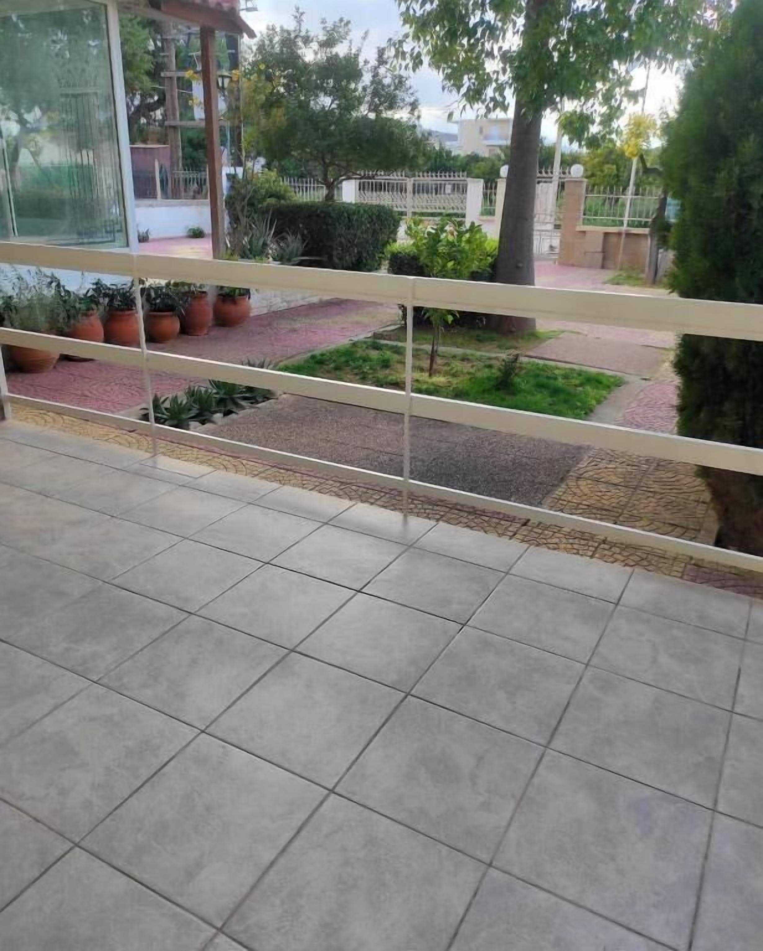 Un balcón con suelo de baldosas, plantas en macetas y vistas a una zona vallada con árboles y un edificio al fondo.