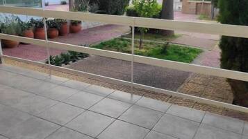 Un balcón con suelo de baldosas, plantas en macetas y vistas a una zona vallada con árboles y un edificio al fondo.