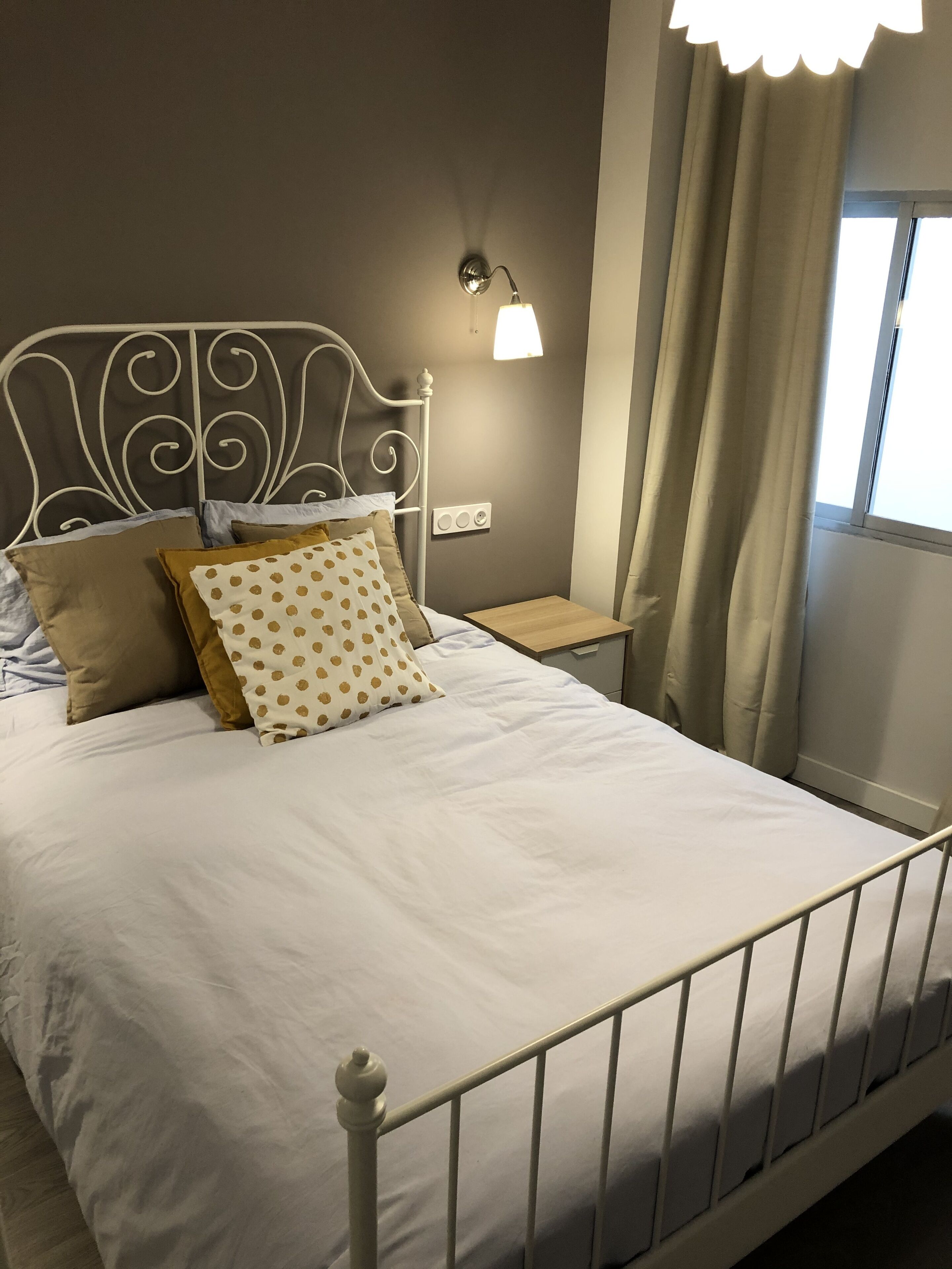 A neatly made bed with white bedding and decorative pillows, a bedside table with a lamp, and a window with curtains.