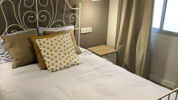 A neatly made bed with white bedding and decorative pillows, a bedside table with a lamp, and a window with curtains.