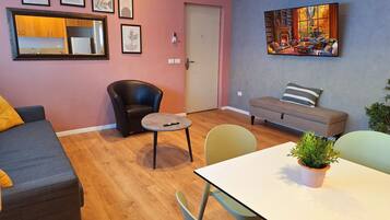Uma sala de estar com sofá, uma poltrona preta, uma mesa redonda pequena, uma mesa de jantar com cadeiras e um quadro na parede.