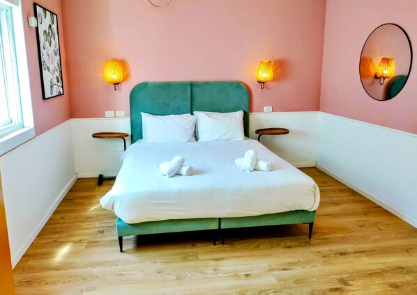 Een netjes opgemaakt bed met witte lakens en opgerolde handdoeken, een blauwgroen hoofdbord en twee bedlampjes.