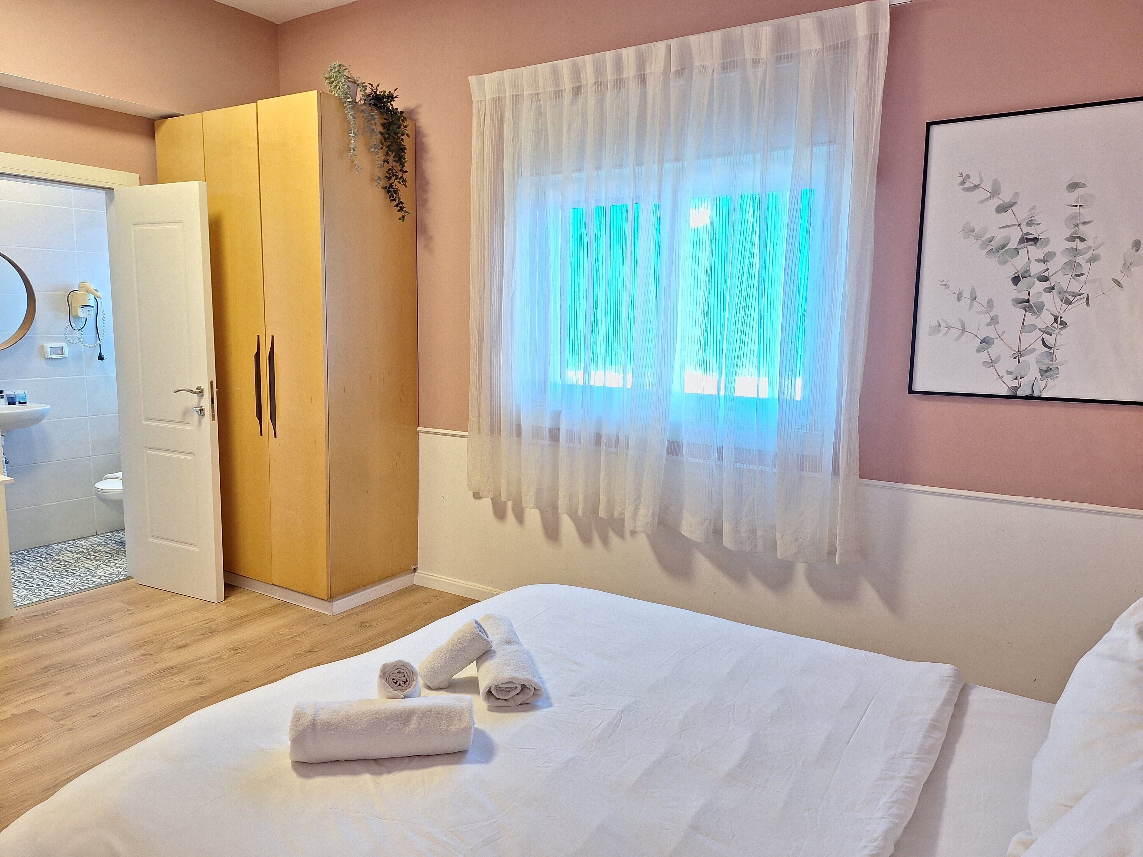 A bedroom with a bed, a wardrobe, a bathroom visible through an open door, and a window with sheer curtains.