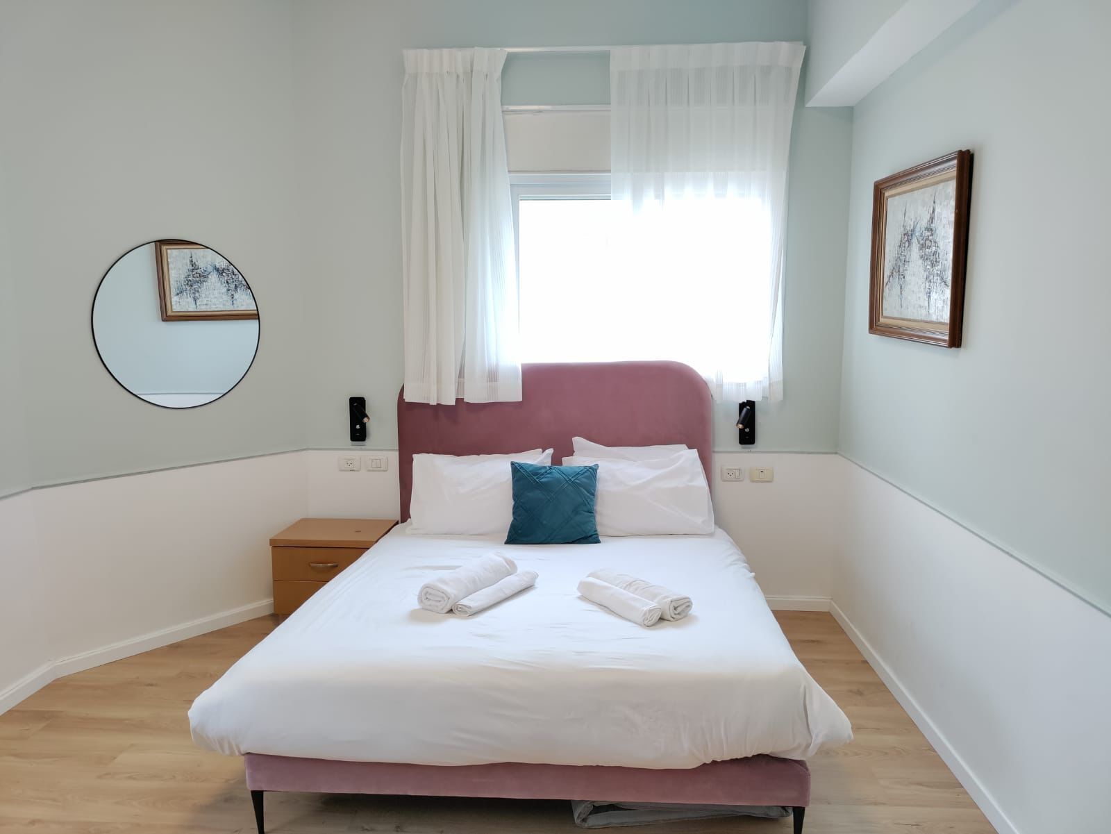 Una cama bien hecha con sábanas blancas y dos toallas enrolladas, un cabecero rosa, una mesita de noche con una lámpara y una ventana con cortinas de encaje.