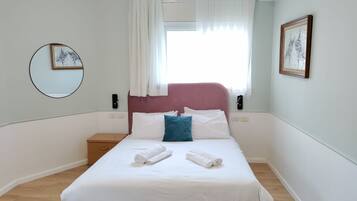 Una cama bien hecha con sábanas blancas y dos toallas enrolladas, un cabecero rosa, una mesita de noche con una lámpara y una ventana con cortinas de encaje.