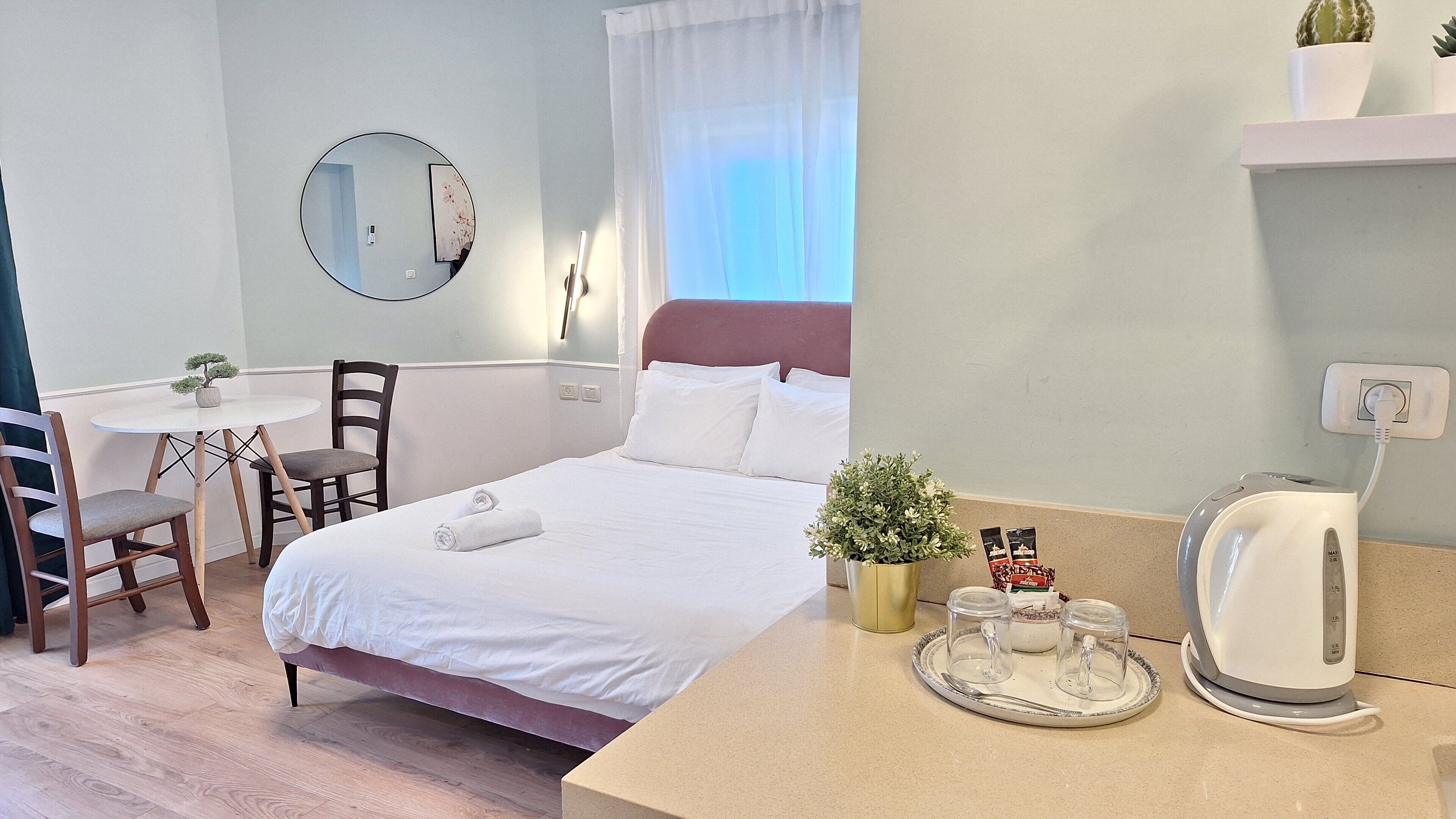 A hotel room with a bed, a round mirror, a small table with a chair, a potted plant, and a kettle on the counter.