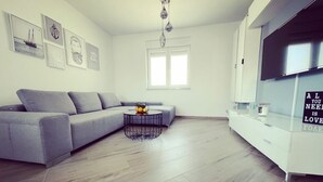 Un salon moderne avec un canapé gris, une table basse ronde sur laquelle sont disposés des fruits, un téléviseur fixé au mur et une grande fenêtre.