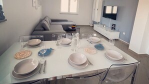 Un espace de salle à manger moderne avec une longue table dressée pour six personnes, des chaises transparentes et une cuisine en arrière-plan.