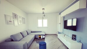 Un salon moderne avec un canapé d’angle gris, une table basse ronde, un pouf bleu, un téléviseur à écran plat fixé au mur et une fenêtre laissant entrer la lumière naturelle.