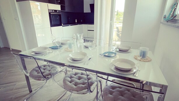 Une cuisine moderne avec des meubles blancs, une table à manger pour quatre personnes et une grande fenêtre qui laisse entrer la lumière naturelle.