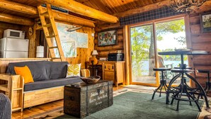 A cozy cabin interior with a wooden ceiling, a sofa with cushions, a dining table, a refrigerator, a microwave, a ladder, and a large window with a view of trees. - Beautiful Private Island Getaway-Boat Rental Available! (Tower)