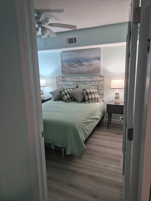 Un dormitorio con una cama, un ventilador de techo, una mesita de noche con lámpara y una obra de arte colgada en la pared.
