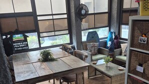 Ein Zimmer mit einem Holztisch, Stühlen, einem Ventilator, einem Bücherregal und Blick durch große Fenster.
