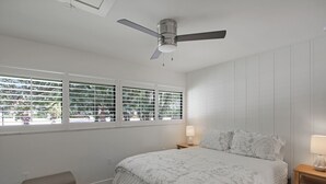 Un dormitorio con cama, ventilador de techo, ventana con persianas, mesita de noche con lámpara y un pequeño banco.
