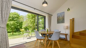 A dining area with a wooden table and chairs, large windows, and a view of a garden. - Bluebell Cottage (Tavistock)