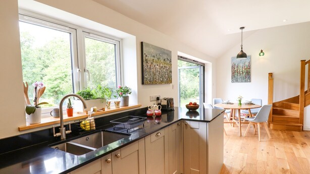A modern kitchen with a black countertop, stainless steel sink, and wooden flooring. There are potted plants on the windowsill and a dining area with a table and chairs. - Bluebell Cottage (Tavistock)