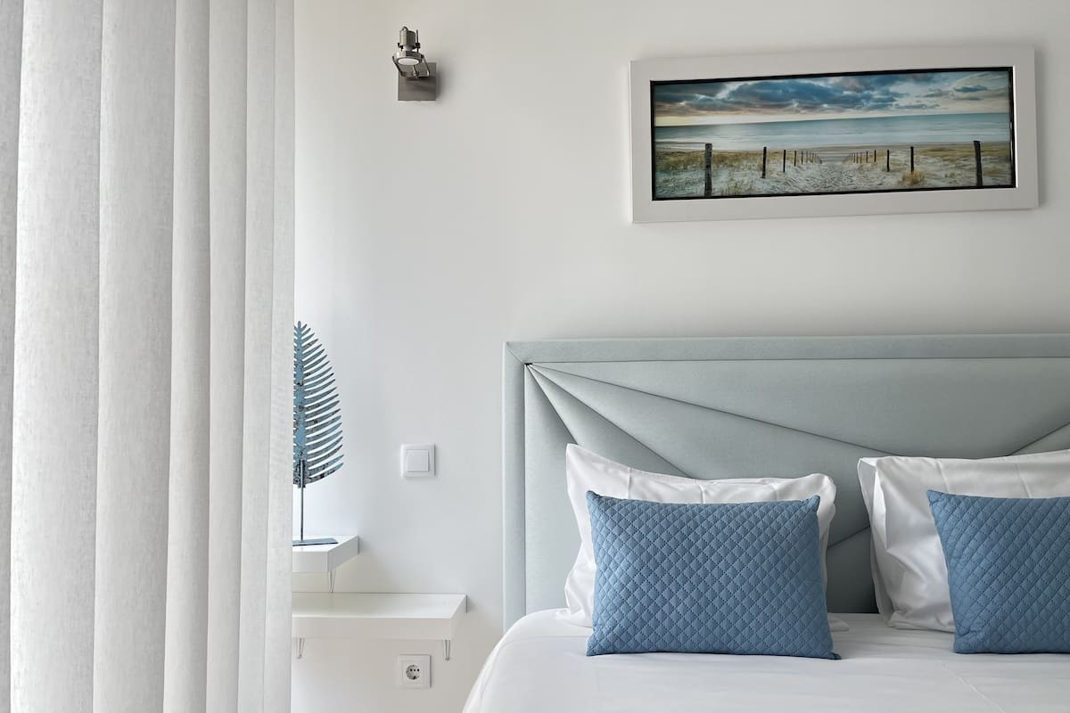 Un dormitorio ordenado con cama, almohadas, mesita de noche, lámpara de pared y un cuadro enmarcados con una escena de playa.