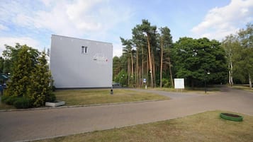 A modern building labeled "Ajaks Hotel" with a paved driveway leading to a parking area, surrounded by trees and greenery.