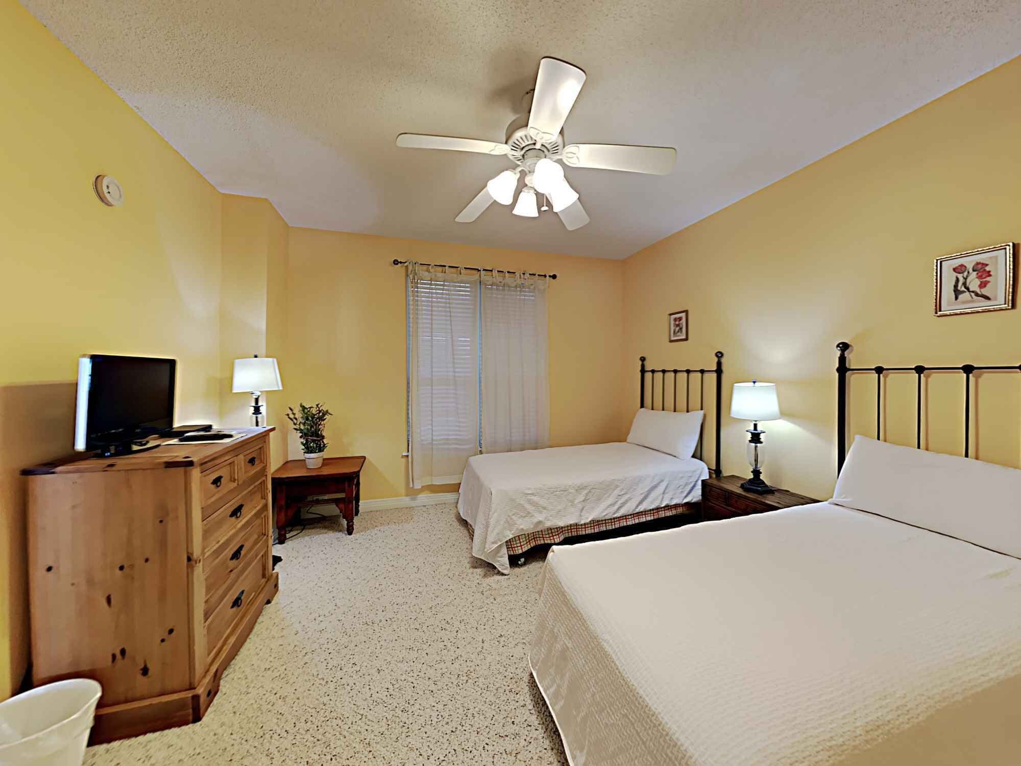 Una habitación de hotel con cama, escritorio con computadora, ventilador de techo y ventana con cortinas.