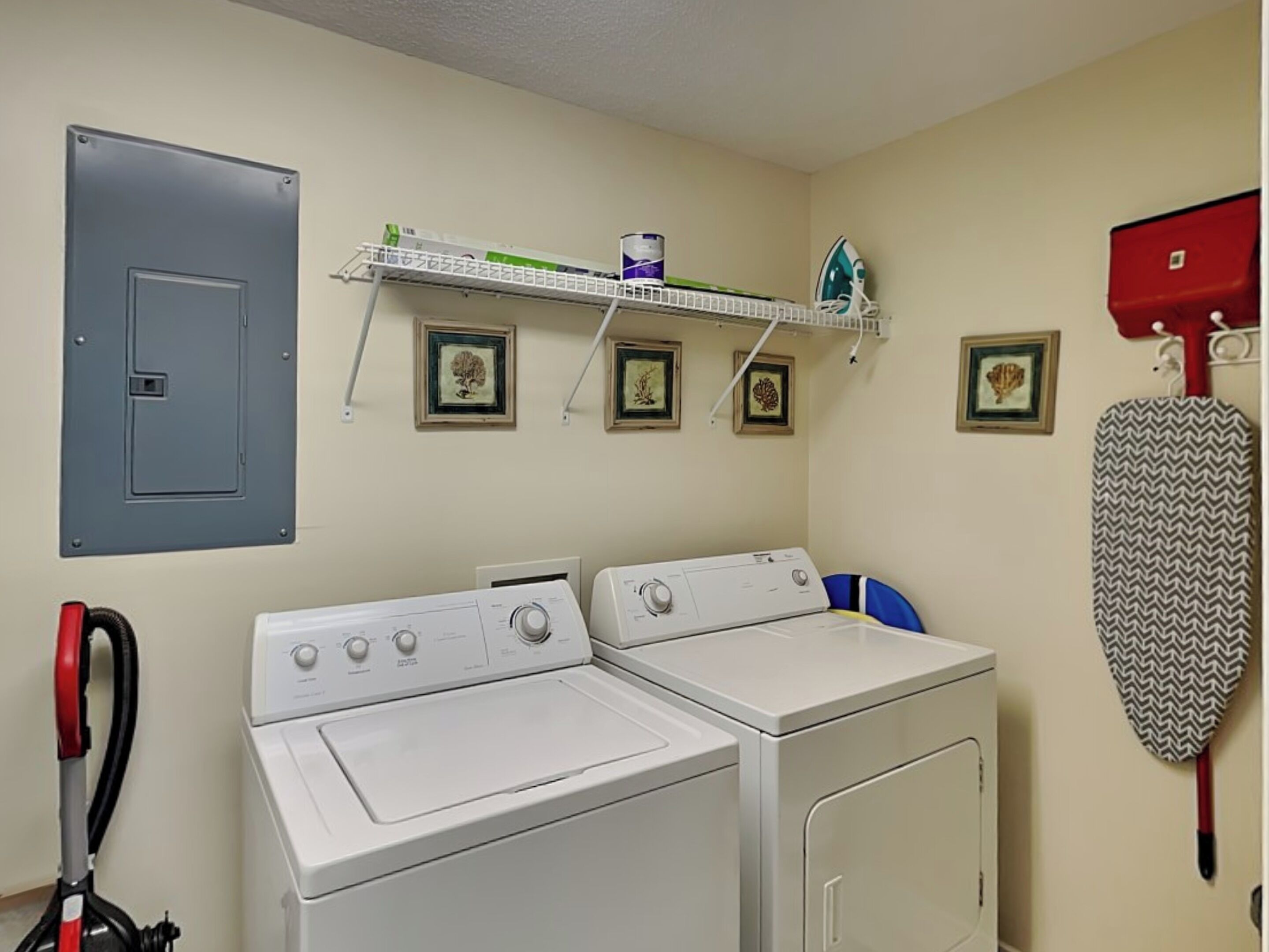 A laundry room with two washing machines, a shelf with framed pictures, a red iron, and a grey utility box.