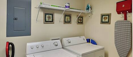 A laundry room with two washing machines, a shelf with framed pictures, a red iron, and a grey utility box.