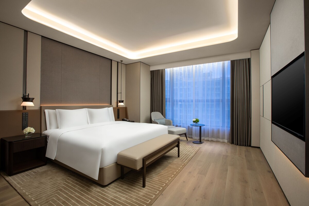 Une chambre d’hôtel moderne avec un grand lit, des tables de chevet, un banc, une petite table avec une plante, une télévision et une grande fenêtre avec des rideaux.
