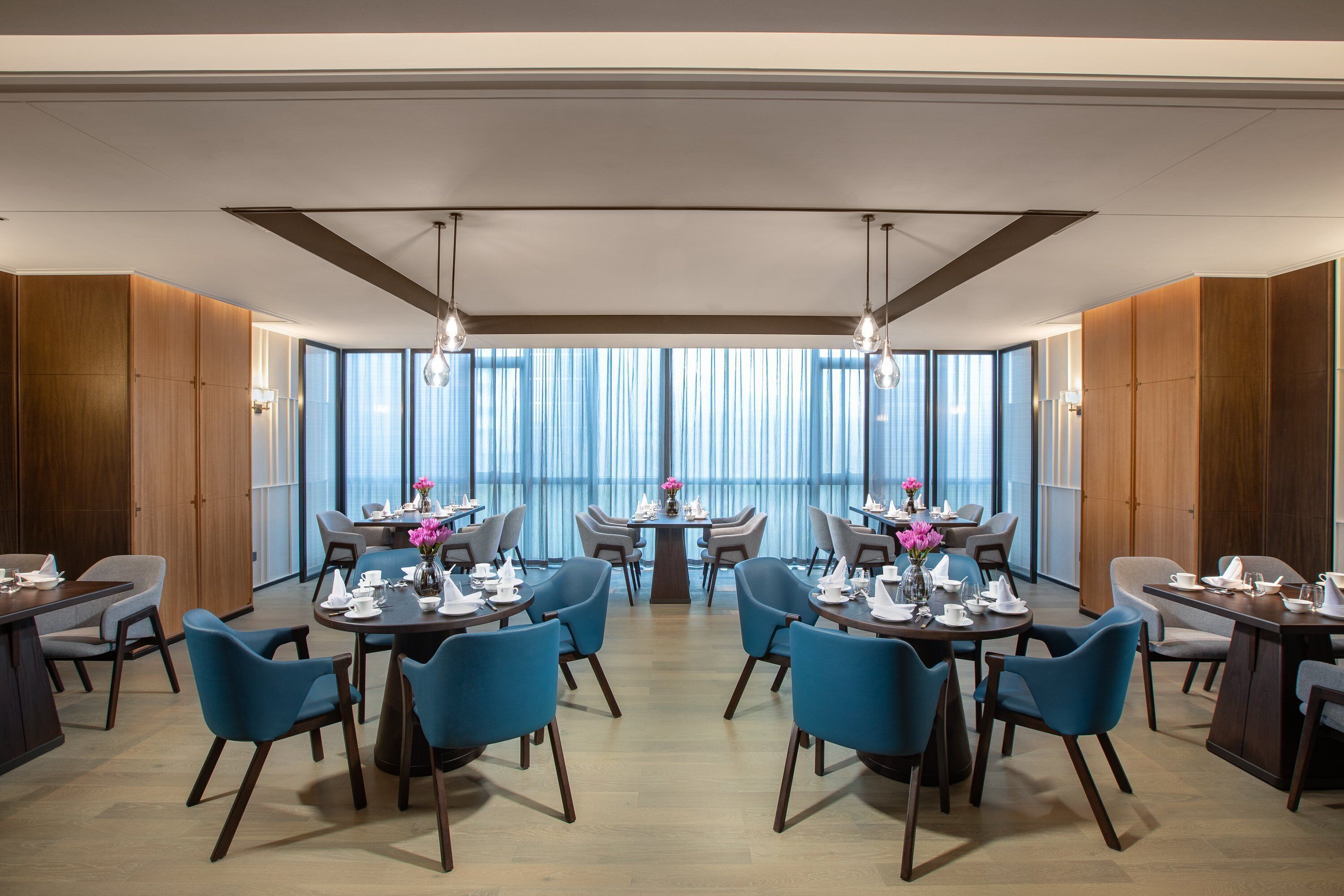 A modern dining area with round tables, blue chairs, and large windows with sheer curtains.