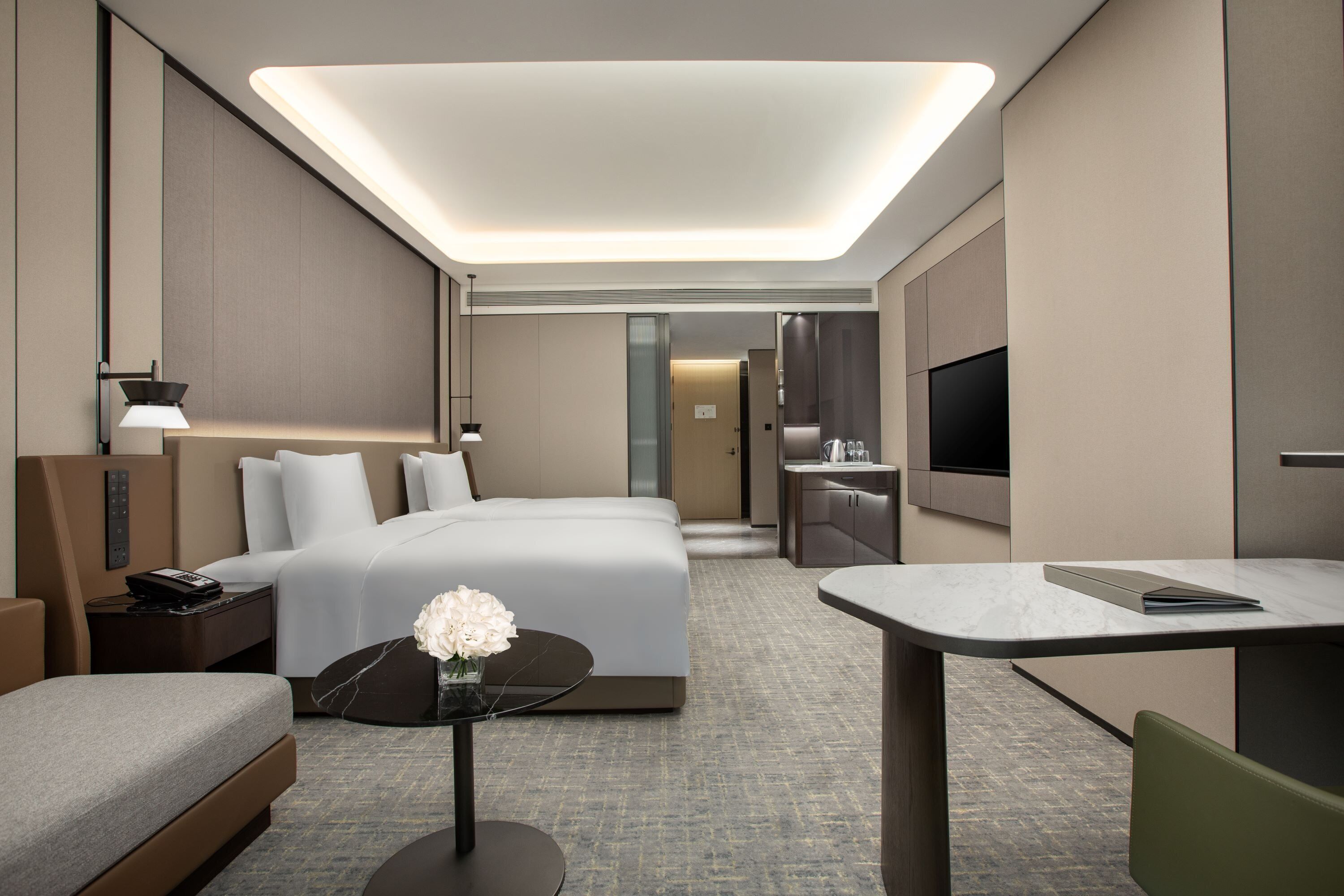 A modern hotel room with a large bed, a desk, a flat-screen TV, and a bathroom visible through a glass door.