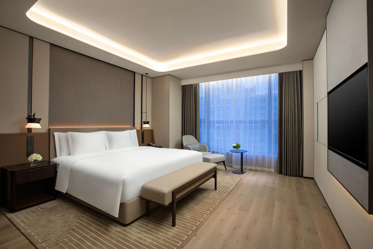 Een moderne hotelkamer met een groot bed, nachtkastjes, een bankje, een klein tafeltje met een plant, een televisie en een groot raam met gordijnen.