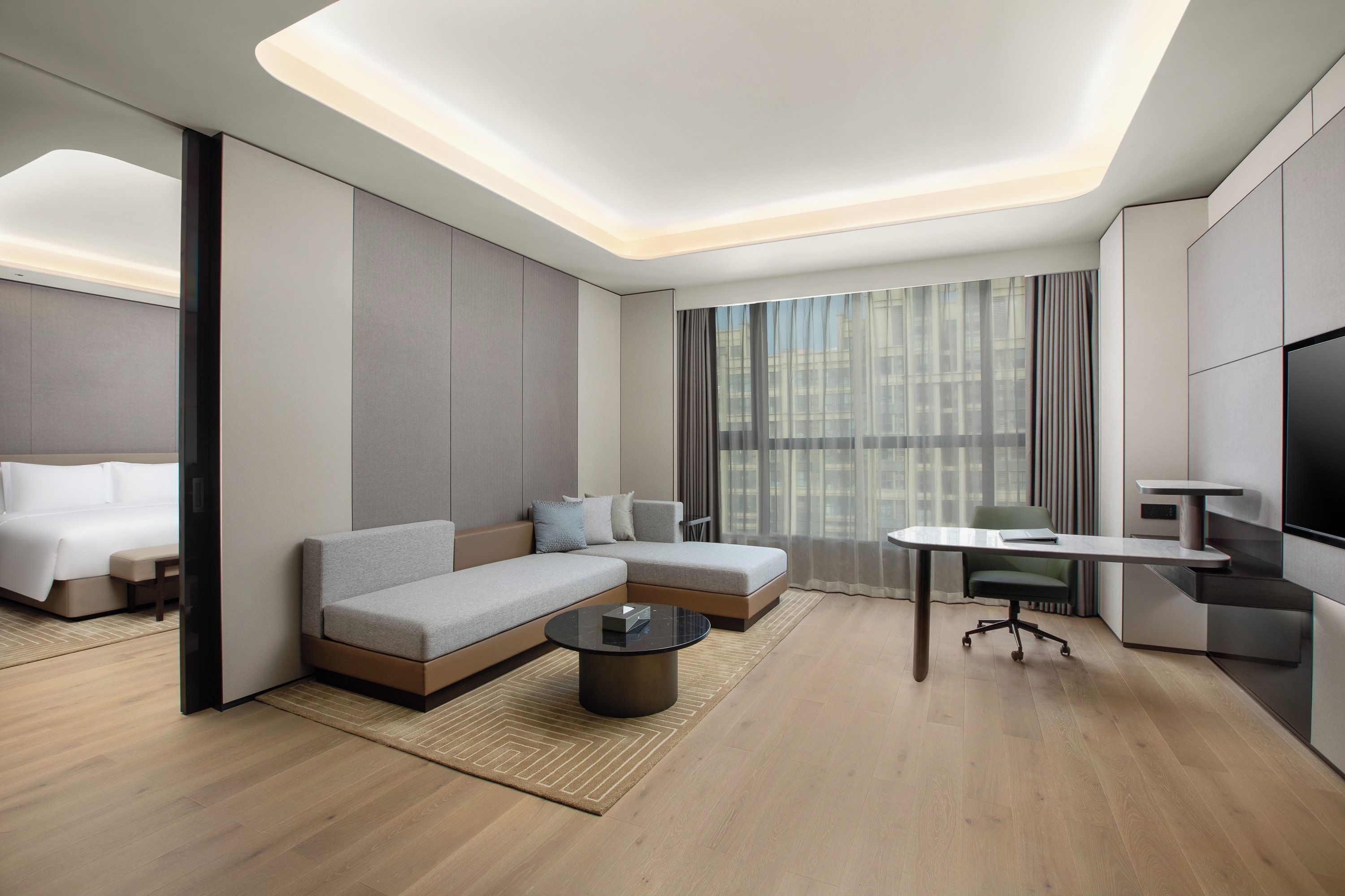 Ein modernes Hotelzimmer mit Sofa, einem kleinen Tisch, einem Schreibtisch mit Stuhl, einem Bett und einem großen Fenster mit Vorhängen.