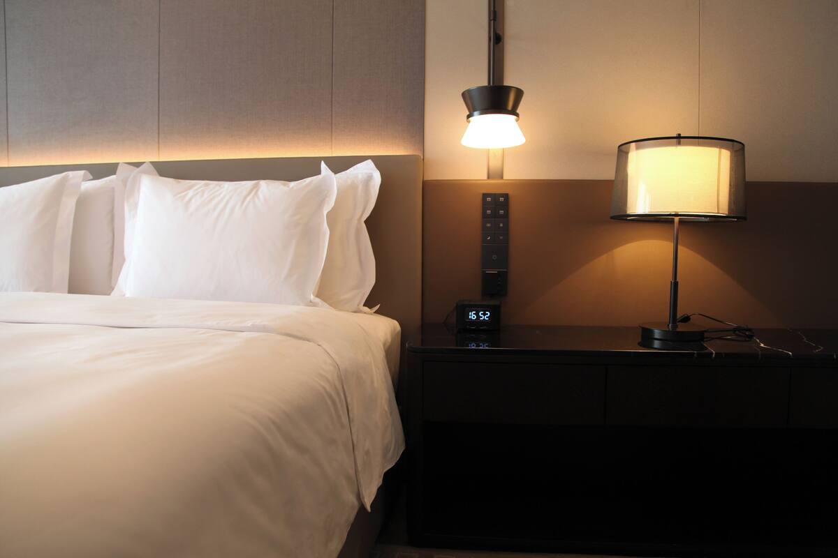 Een keurig opgemaakt bed met witte lakens, een nachtkastje met een lamp en een digitale klok waarop de tijd staat.