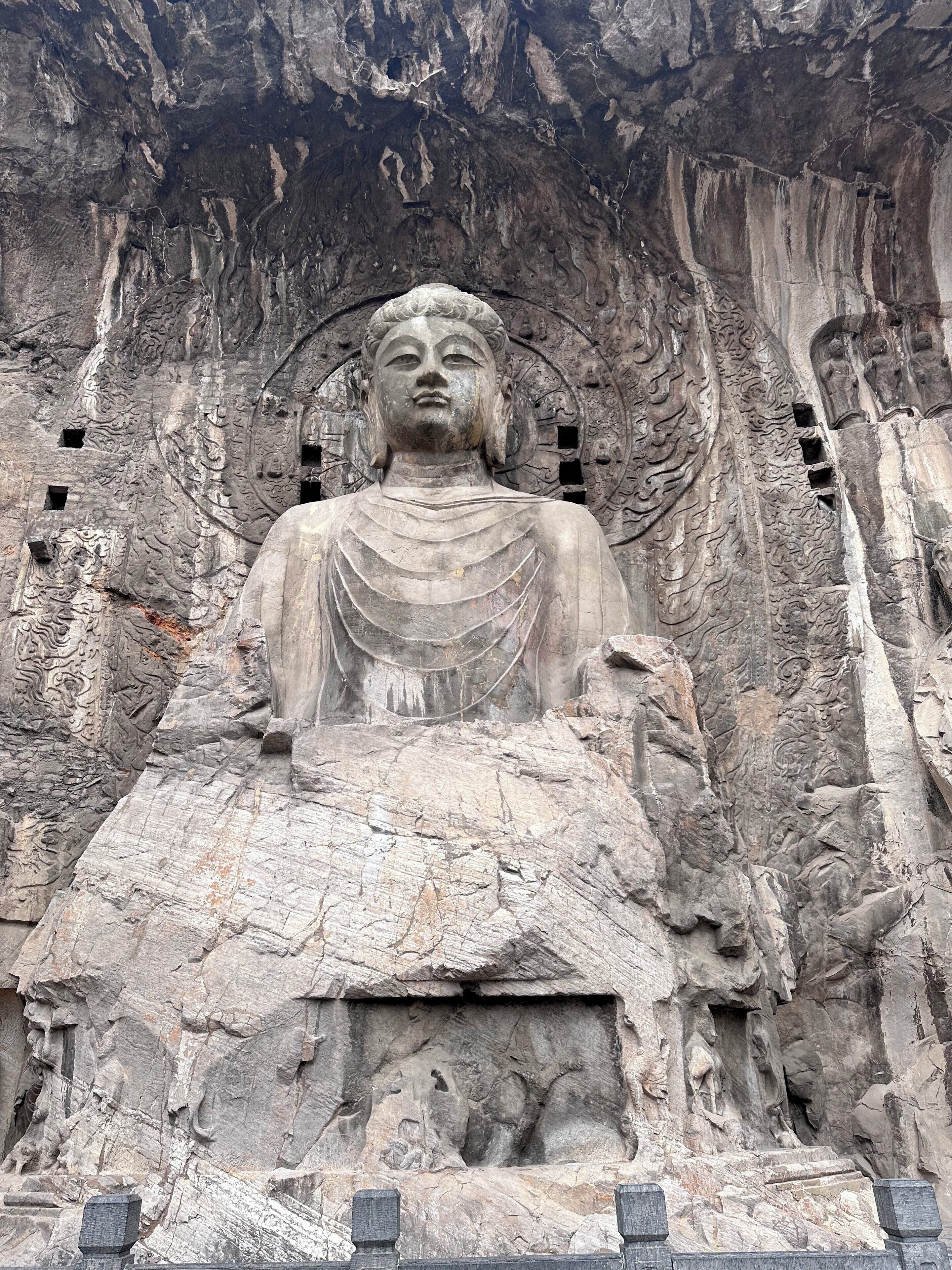 A large stone Buddha statue with intricate carvings and a serene expression, set against a rugged rock face.