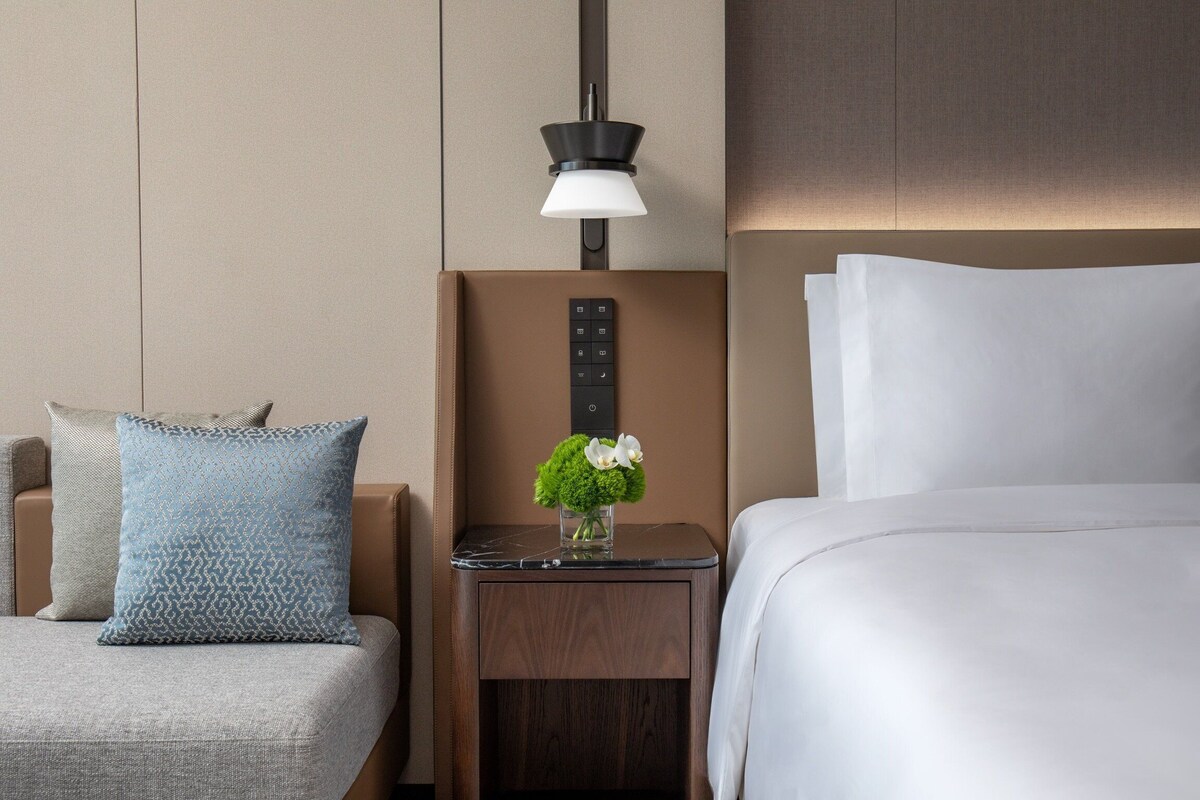 Une chambre d’hôtel avec un lit, une table de chevet, une lampe, un vase avec des fleurs et un oreiller.
