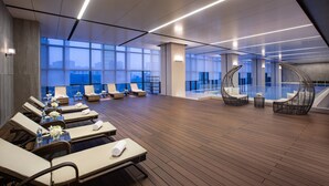Een ruime lounge met ligbedden en tafels, grote ramen met uitzicht over de stad, en moderne verlichting.