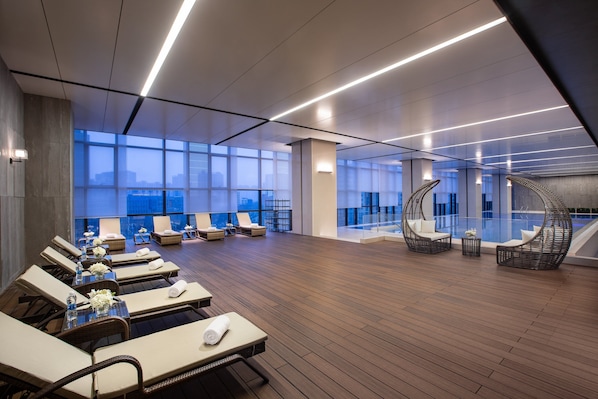 Een ruime lounge met ligbedden en tafels, grote ramen met uitzicht over de stad, en moderne verlichting.