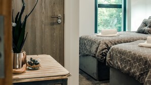 A bedroom with two beds, a wooden door, a window with a view, and a small table with a plant. - Hayy Collection Townhouse Living at Cliff Oaks (Leeds)