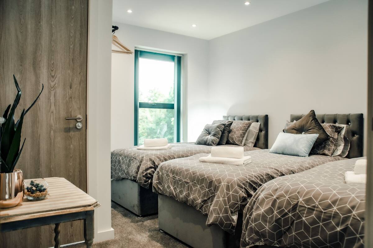 A hotel room with two beds, a wooden door, a small table with a plant, and a window with a view of greenery.