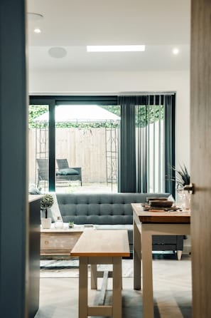 A modern living room with a grey sofa, wooden coffee table, and a dining table. There is a view of a wooden fence and greenery outside.