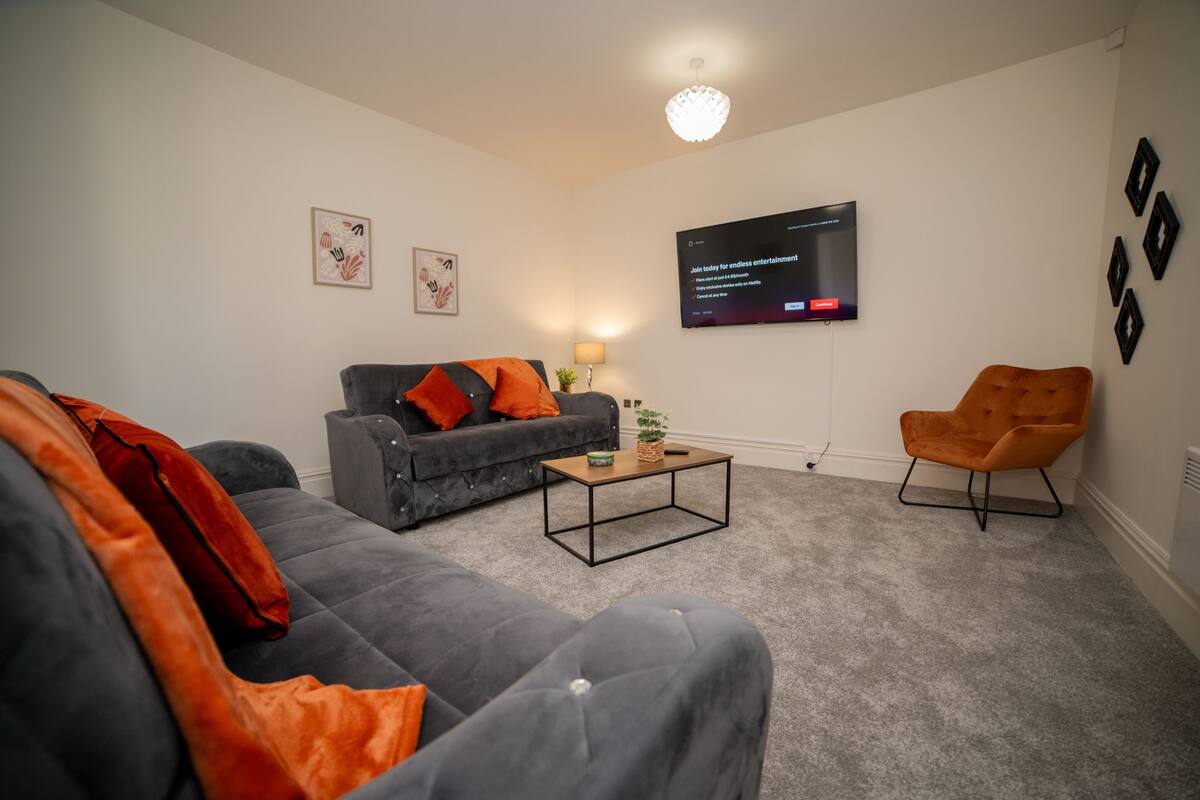 A modern living room with a grey sofa, an orange armchair, and a flat-screen TV mounted on the wall.